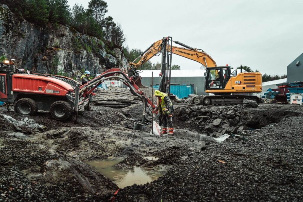 Tom menn i arbeidsklær jobber ute i regnet ved siden av en borerigg. Gravemaskin i bakgrunnen.