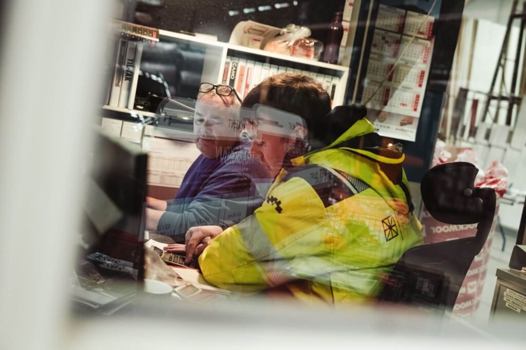 To menn sitter og arbeider inne på et kontor i arbeidsklær